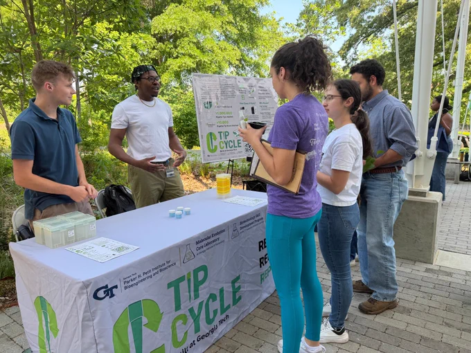 People gathered at an outdoor Georgia Tech “TIP Cycle” information table, discussing lab tip recycling and sustainability.
