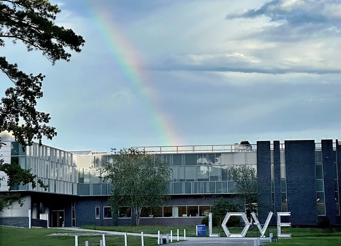 The image shows a large building with reflective windows under a cloudy sky, featuring a distinct rainbow. In the foreground, there's a sculpture spelling out "LOVE" made of white letters. 