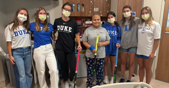 A group of people in a hospital room, all wearing masks. Most are wearing Duke t-shirts. One person in the center is in hospital attire.