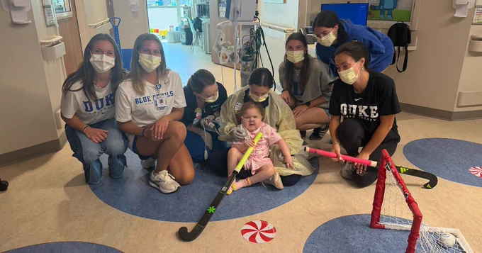 A group of individuals in Duke University apparel are gathered around a young child sitting on the floor, holding toy hockey sticks. They are in a room with playful, colorful decorations. The individuals are wearing face masks.