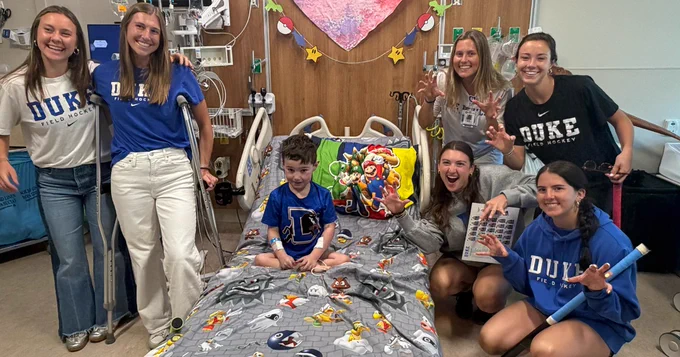 A group of people in Duke field hockey shirts are gathered around a hospital bed with a young child sitting on it, decorated with Super Mario-themed bedding. The room is adorned with a colorful heart-shaped decoration and other hanging ornaments. Everyone is smiling and appears to be enjoying the visit.