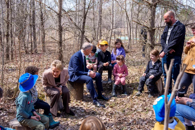 Prime Minister Mark Carney and Diana Fox Carney are seated on tree trunks in a nature park, surrounded by children.