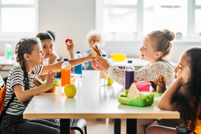 Five children sit at a table in a bright room, eating lunch. The table has colorful lunch boxes, water bottles, and fruit. Two children are smiling and passing food.