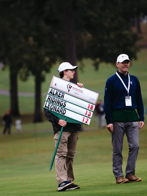 A person stands on a golf course holding a leaderboard sign displaying the names Alker, Ridings, and Leonard with corresponding scores. Another individual stands nearby, both wearing caps. The background features grass and trees.