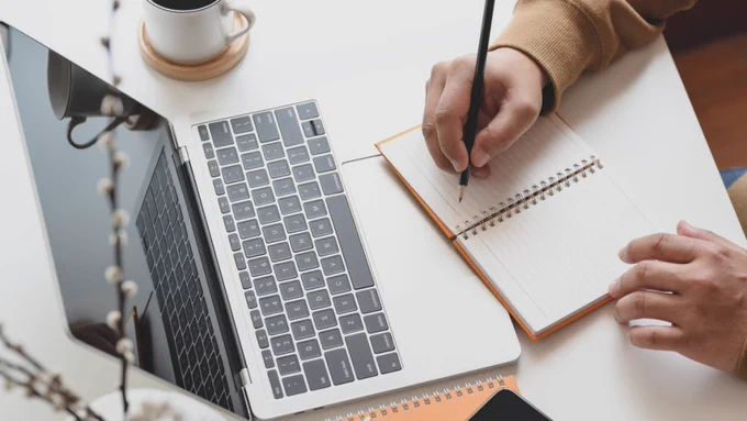 A person writes in an orange notepad next to a laptop and coffee mug on a white desk. A cell phone and another notebook are visible at the bottom of the frame.
