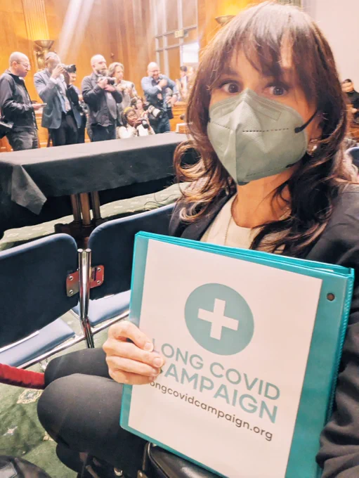 Wheelchair user Meighan at today's the Senate HELP Committee hearing. wearing a N95 mask due to being immune compromised, holding a binder for the Long COVID Campaign, while a crowd of press photographers gather to film Secretary Kennedy's arrival to testify on the FY2027 HHS budget request.
