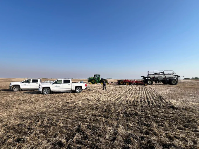 A wide field under a clear blue sky is shown. Two white pickup trucks with a Doulbe Diamond Farm Supply logo on the side are parked on the left. In the center, a green John Deere tractor is attached to farming equipment. A person is walking nearby. On the right, a large piece of machinery is visible. The ground is covered with dry, harvested crops.