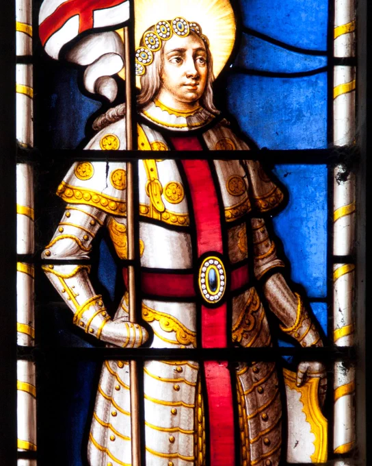 Stained glass window depicting St George in ornate armor holding a flag with the cross of St. George. St George is adorned with a halo and stands against a blue background.