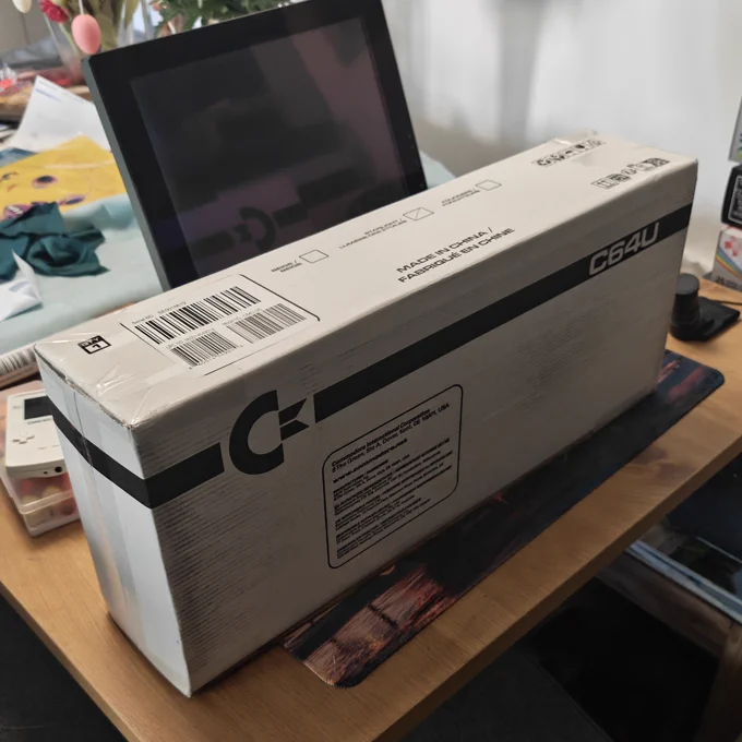 Das Foto Zeigt die Umverpackung des Commodore 64 Ultimate. Sie ist weiß mit schwarzer Schrift, das Commodore Logo ist prominent auf der Schachtel.
Diese liegt auf einem Holztisch, dahinter ein kleiner LCD Monitor. 