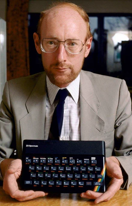A man in a suit and glasses holds a ZX Spectrum computer, showcasing it proudly. The scene conveys a sense of innovation and nostalgia.