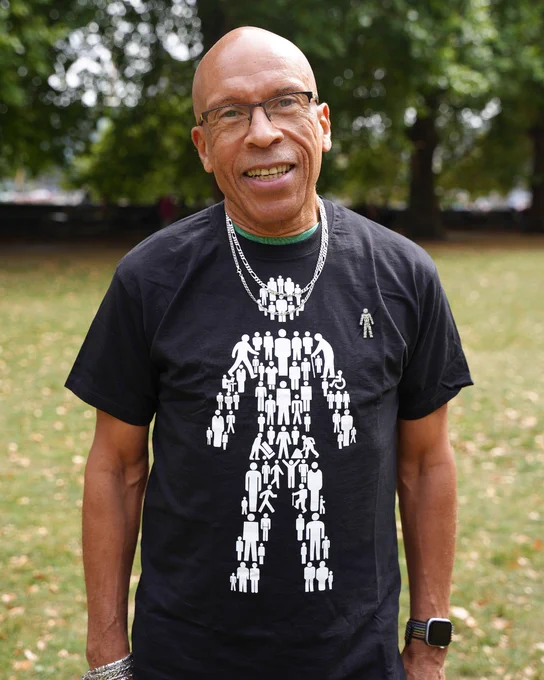 Des stands outdoors in a park, wearing glasses and a Prostate Cancer UK Man of Men t-shirt. The background has green trees and grass.