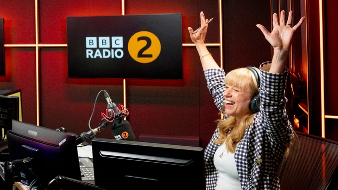 A woman sits in a radio studio smiling, her arms raised in the air in celebration