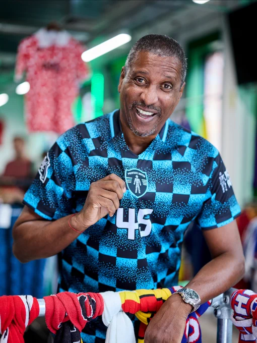 Viv Anderson stands smiling in a classic football shop wearing a Prostate United football shirt, holding up the badge to the camera. 