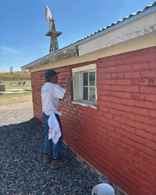 Man painting the exterior red brick wall of a small building under a clear blue sky