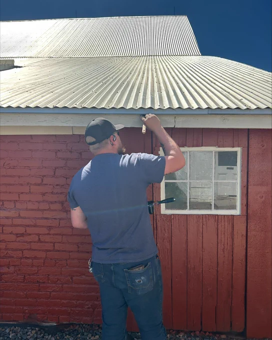 Man in a cap painting a red barn wall under a corrugated metal roof on a sunny day