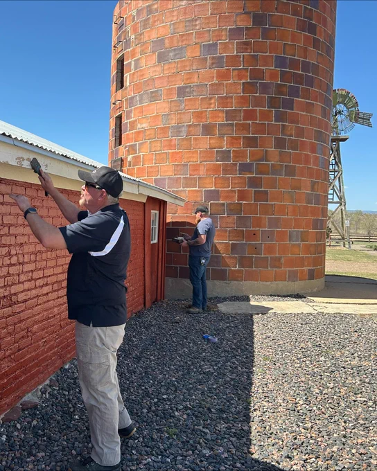 Two men painting a wall outside near a large cylindrical brick silo under a clear blue sky
