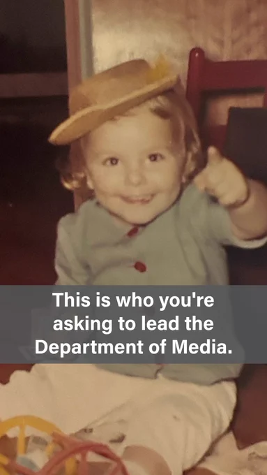 A vintage childhood photo of a smiling toddler wearing a light blue jacket with red buttons and a tan/brown hat tilted on their head, pointing a finger toward the camera with a playful expression. Text overlay reads: "This is who you're asking to lead the Department of Media."