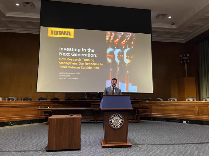 A man speaks in front of a podium with a screen behind him that says: Investing in the next generation: How research training strengthens our response to rural veteran suicide risk.