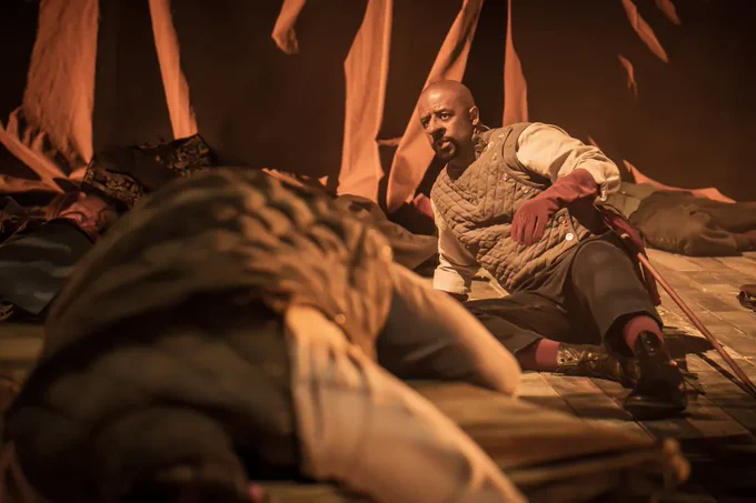 A photo showing a man in doublet and gloves lying on a stage 