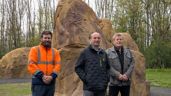Three men standing in front of large artificial rock formations in an outdoor setting. The man on the left is wearing a bright orange high-visibility jacket, while the other two are dressed in darker jackets. The background includes tall, leafless trees and green grass.