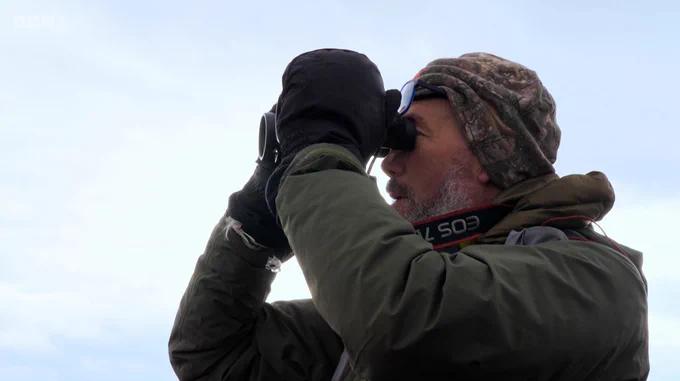 Dr Carlos Bedson using binoculars against a pale blue sky.