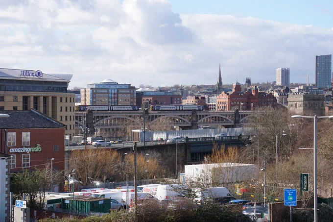 A cityscape featuring the Hilton hotel, various buildings, and distant architectural structures. The image includes a bridge with multiple arches with two Northern trains travelling across, and a skyline with a prominent spire. In the foreground, there is a collection of parked vans surrounded by trees.