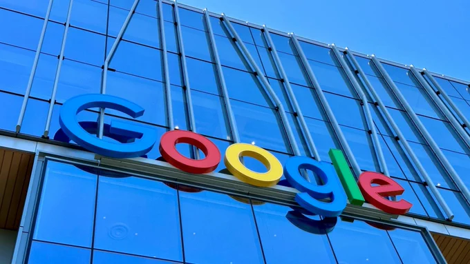 The colorful Google logo mounted on the glass facade of a Google office building against a blue sky.