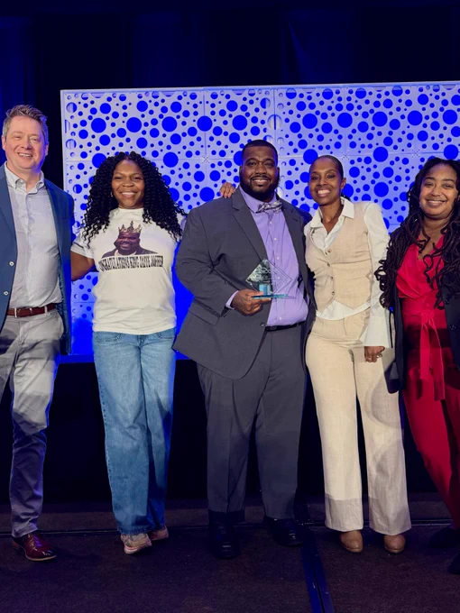 Jeromy standing in the middle of the image holding his award while being surrounded by SJCS staff.