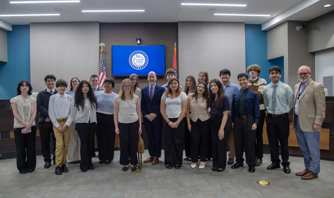 A group of people gathered for an event in an auditorium with an American flag and a logo on the wall behind them.