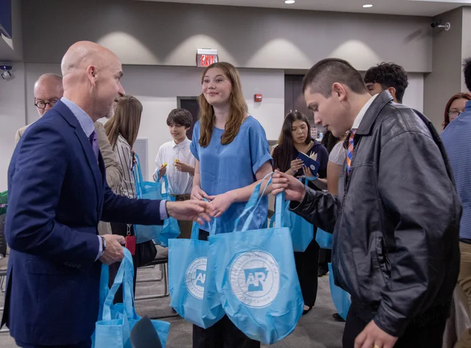 Several people standing in a hallway, a man handing out blue bags with a logo on them to a group of students.