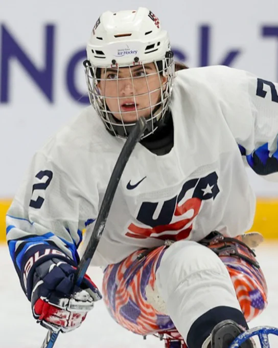 A sled hockey player in a white USA jersey and helmet, gripping a stick, focused on the game.