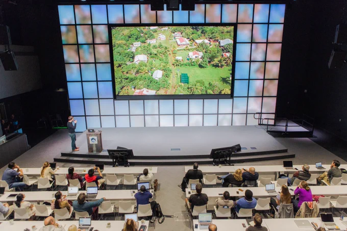 People seated at tables watch a person presenting on a stage, where a large screen shows an image of residential homes surrounded by greenery.