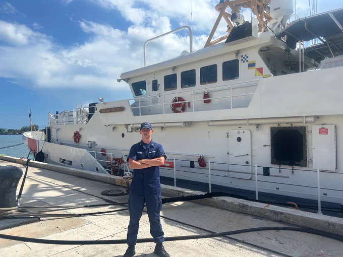 A person in a coast guard uniform stands on a concrete pier with a white ship in the background