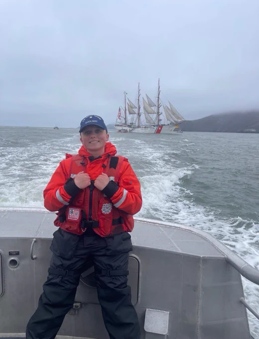 A woman in a coast guard uniform sits on the back of a boat with a tall ship in the background