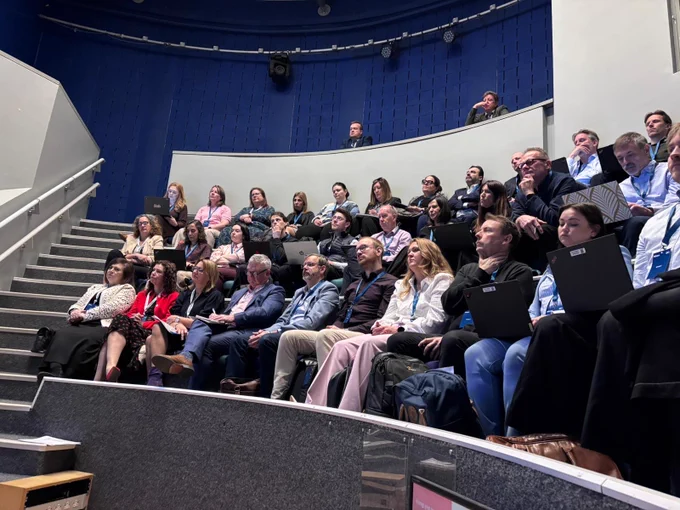 A group of people seated in a tiered lecture hall, attentively listening to a presentation. Some attendees have laptops open on their laps. The room has a modern design with blue walls and bright lighting.