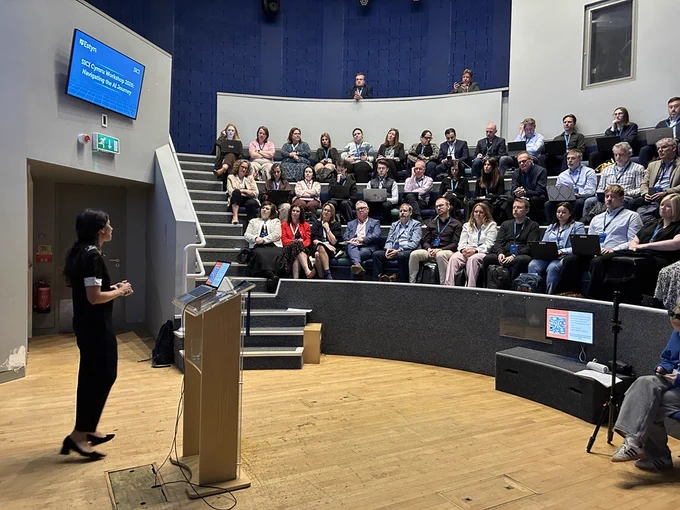 A person is presenting in a modern lecture hall with a curved seating arrangement. Attendees are seated, attentively listening to the presentation.