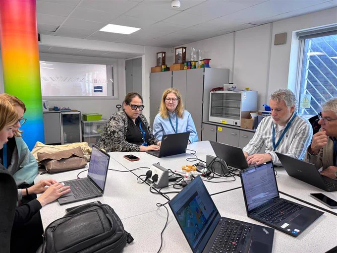A group of individuals are seated around a table, each working on laptops. The room has a colorful pillar and various cabinets. Several laptops display spreadsheets and other documents. The setting appears to be a collaborative workspace or meeting room.