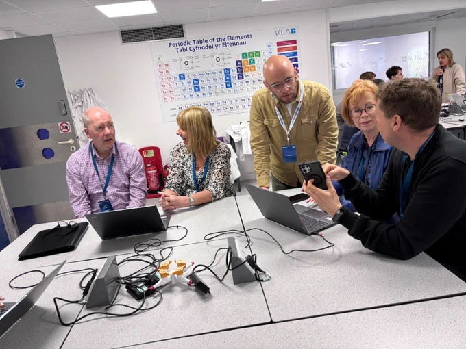 A group of people is gathered around a table with laptops and cables. In the background, a periodic table of elements is displayed on the wall. The setting appears to be a classroom or workshop environment.