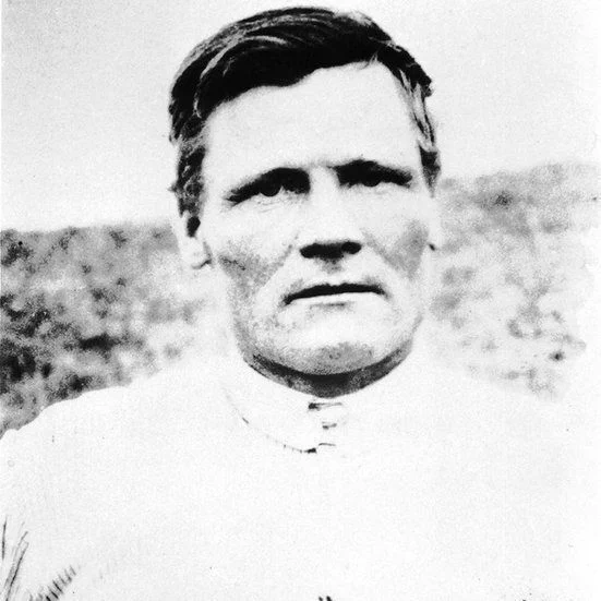 Tom Sukanen faces the camera in a black-and-white portrait, wearing a light-coloured shirt with a high collar. His hair is combed to the side, and he has a serious expression. The background is outdoors and softly blurred.
