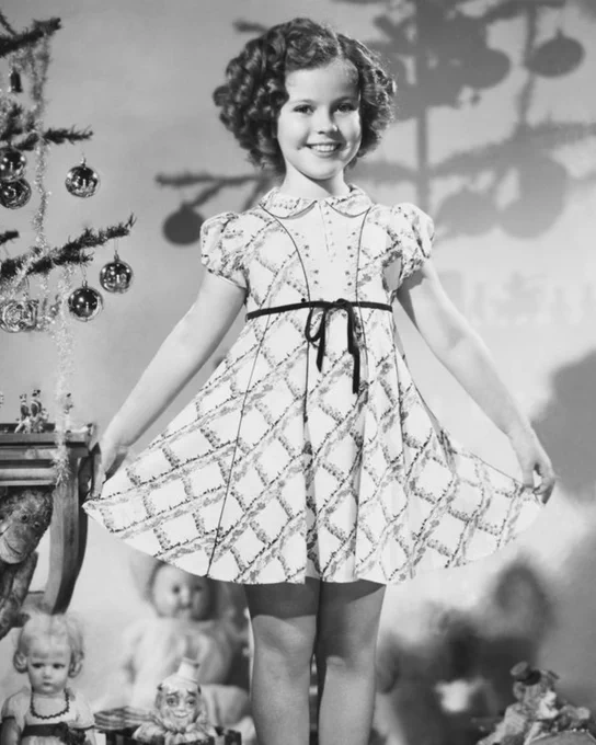 Shirley Temple as a child, smiling and holding out the skirt of her dress. 