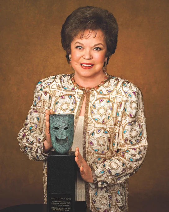 Shirley Temple smiling and resting her hands on her SAG Life Achievement Award. 