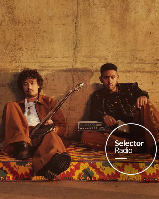 Two musicians from the duo ZENA sit against a sunlit concrete wall, one holding an electric guitar and the other resting a hand on a compact music device, both dressed in stylish coordinated brown outfits on a vibrant patterned mattress. Warm golden-hour lighting casts long shadows. The Selector Radio logo appears in a white circle in the bottom right corner.