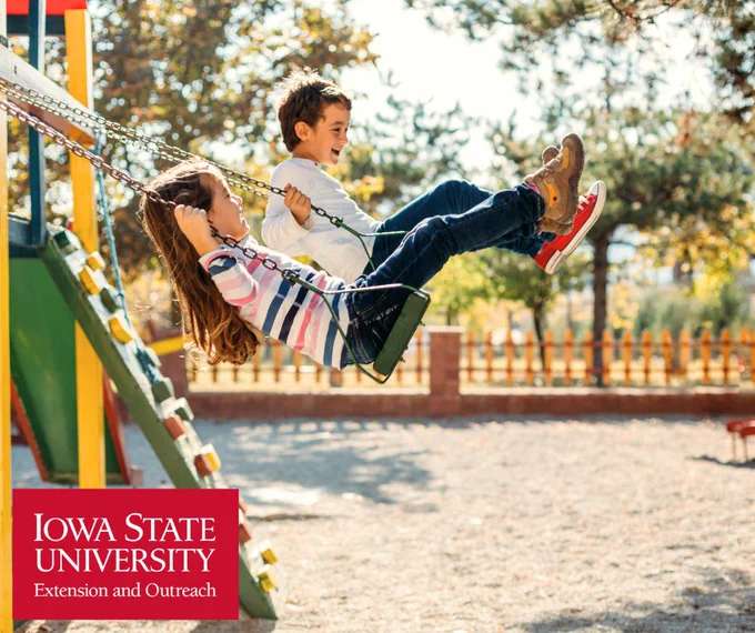 Two children swinging side by side on a playground swing set on a sunny day.