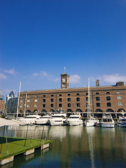 Waterfront scene: several yachts docked in front of a brick building with a clock tower under a blue sky. Reflections shimmer on the water's surface, adding depth to the urban landscape.