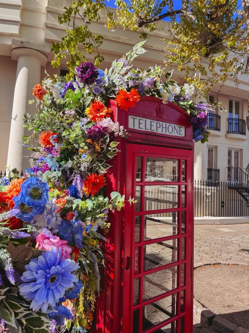 A vibrant display of colorful flowers climbs up a classic red telephone booth with \"TELEPHONE\" written on the front. The booth stands on a brick street next to the main campus building. Sunlight filters through the leaves.