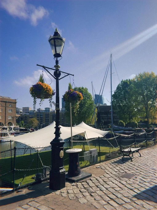 A waterfront scene on a sunny day features a classic black lamppost adorned with hanging flower baskets. A white tent and sailboats are visible in the background. A bench sits along a brick path by a black chain-link fence.