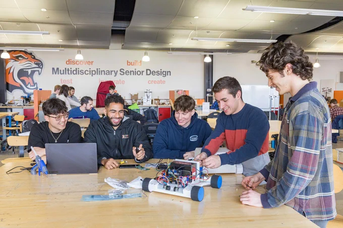A team of five students sit at a large table workstation in a large open workshop. They are working on a small wheeled robot with exposed wires while using a laptop and remote control. 
