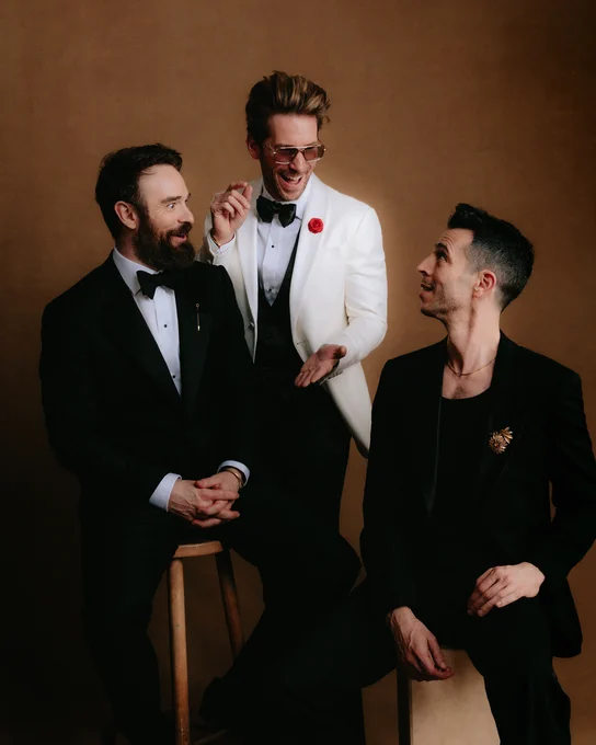 Charlie Cox (left, seated) Troy Baker (middle, standing) and Shai Matheson (right, seated) pose for a portrait. Shai looks at Troy, who laughs. Charlie has an amused expression. All three wear formal suits.