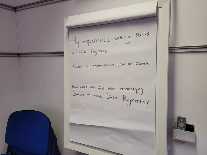 Flip chart on a stand in a room with stacked blue chairs showing handwritten text reading “My experience getting started with Direct Payments”, “Support and communication from the council”, and “How would you feel about encouraging someone to have Direct Payments?” with wall fittings in the background.