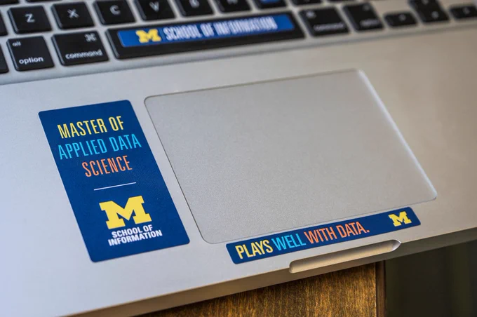 Close-up of a laptop touchpad and keyboard decorated with UMSI Master of Applied Data Science stickers.